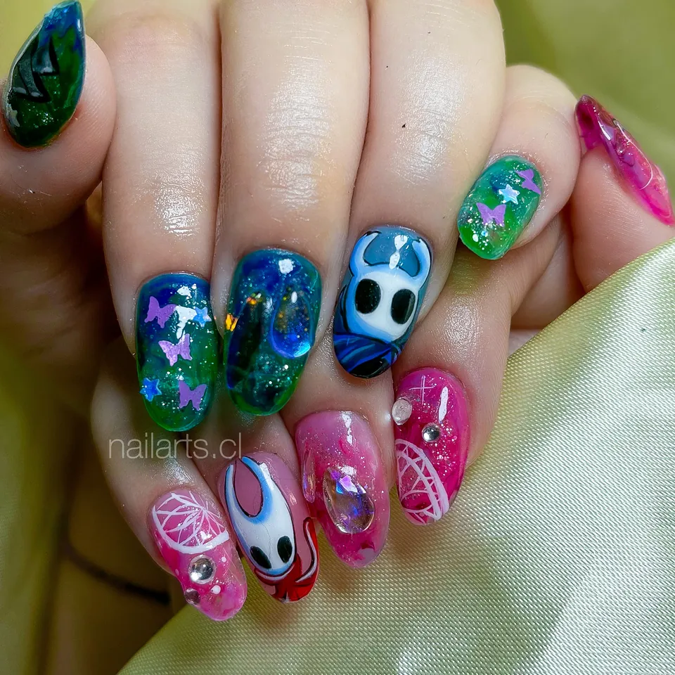 Uñas verdes, azules y rosadas con un personaje de videojuego de máscara blanca y mariposas
